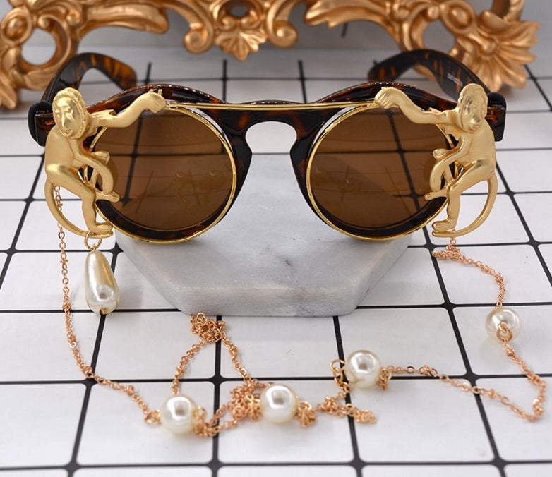 Sunglasses with gold monkey designs and a pearl chain on a checkered surface.