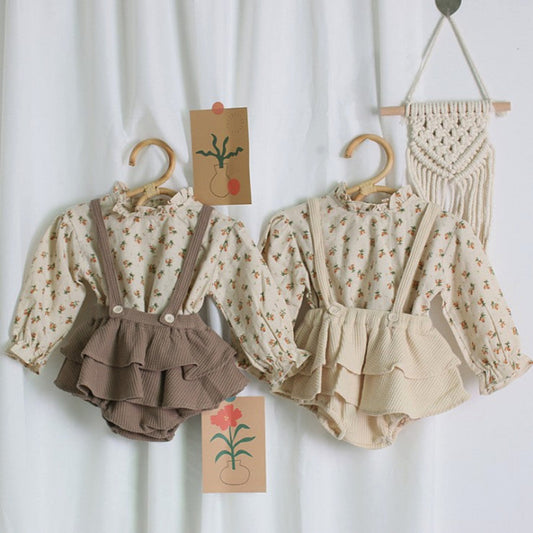 Children's floral blouse with ruffled shorts and suspenders on hangers against a white curtain background.