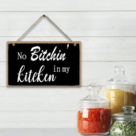 Decorative sign with text 'No Bitchin' in my kitchen' hanging on a white wooden wall with jars of spices below.