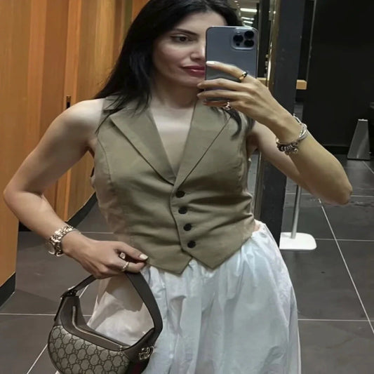 Woman taking a mirror selfie wearing a beige vest and white dress, holding a handbag.