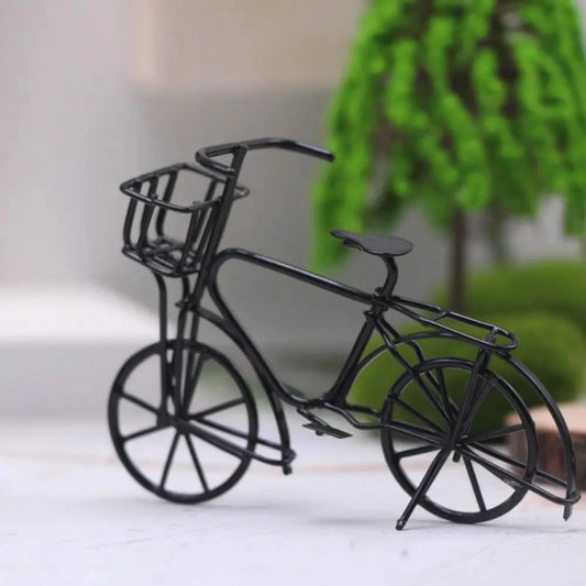 Black metal bicycle model with a basket on a white surface with a blurred green plant in the background