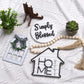 Decorative items including a house-shaped sign, wreath, and 'Simply Blessed' plaque on a textured white surface.
