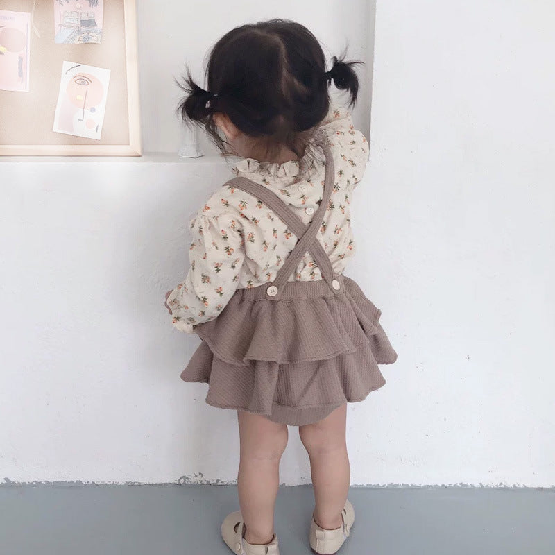 Child wearing a floral blouse and brown ruffled skirt with suspenders against a white wall.
