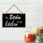 Decorative sign with text 'No Bitchin' in my kitchen' hanging on a white wooden wall with jars of spices below.
