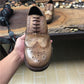 Brown leather shoe held by a hand with a camera and wooden surface in the background