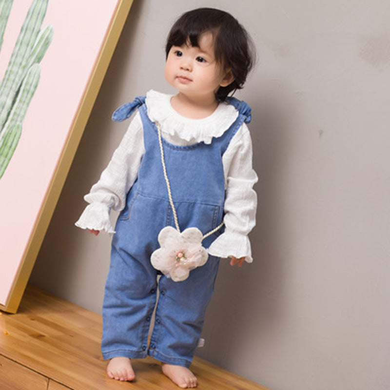 Child wearing a blue denim dress overalls with a white shirt, standing against a plain wall.