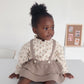 Child wearing a floral blouse and brown ruffled skirt sitting on a white surface.