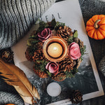 Decorative candle holder with flowers and pinecones on a magazine with a pumpkin and feather beside it.