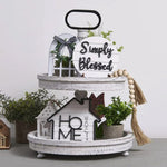 Decorative tiered tray with 'Simply Blessed' and 'Home' signs, plants, and tassels on a light background.