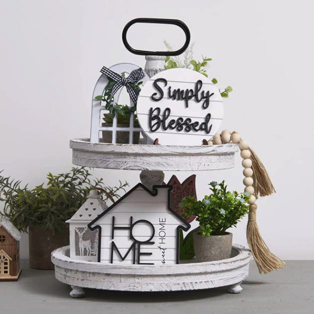 Decorative tiered tray with 'Simply Blessed' and 'Home' signs, plants, and tassels on a light background.
