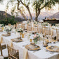 Elegant outdoor wedding reception with tables set for dinner, chairs, and floral arrangements under a tree.