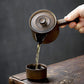 Person pouring liquid from a small pot into another container on a wooden surface with a dark background