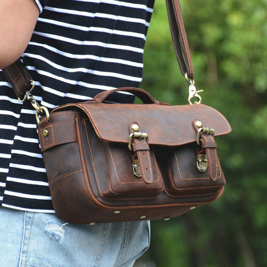 Brown leather satchel bag held by a person wearing a striped shirt and jeans, with a blurred green background.