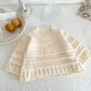 Knitted baby sweater on a white surface with a plate of pastries and a glass in the background.