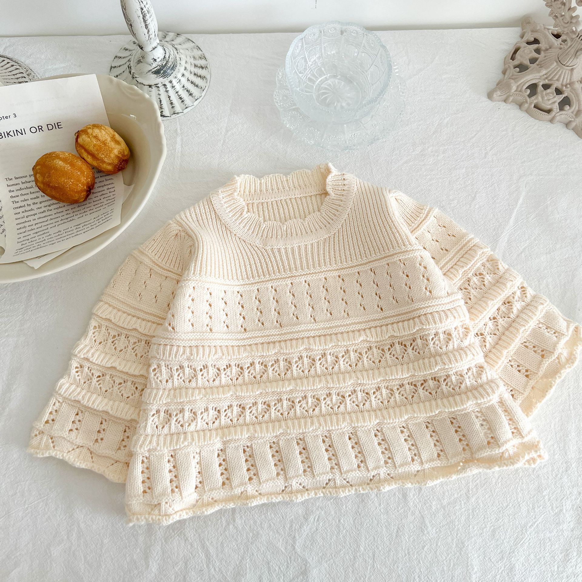 Knitted baby sweater on a white surface with a plate of pastries and a glass in the background.