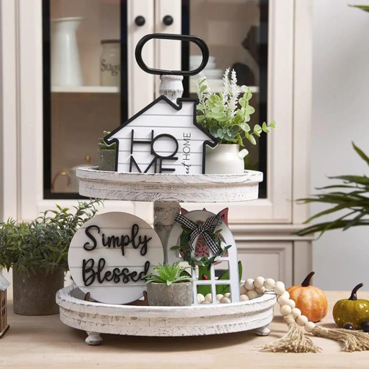 Decorative tiered tray with 'Home' and 'Simply Blessed' signs, pumpkins, and plants on a table.