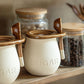 Ceramic sugar jar with wooden lid and spoon on a shelf with glass jars in the background.