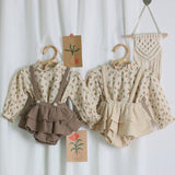 Children's floral blouse with ruffled shorts and suspenders on hangers against a white curtain background.