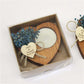 Heart-shaped wooden coaster with dried flowers and a candle, tied with twine and a heart-shaped tag, in a box.