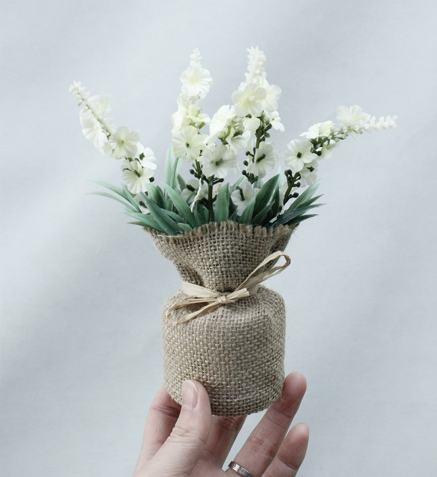 Bouquet of white flowers in a small jar wrapped with burlap and tied with a ribbon, held against a light gray background.