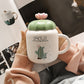 Person holding a cactus-themed mug with a green lid, surrounded by blurred indoor background.