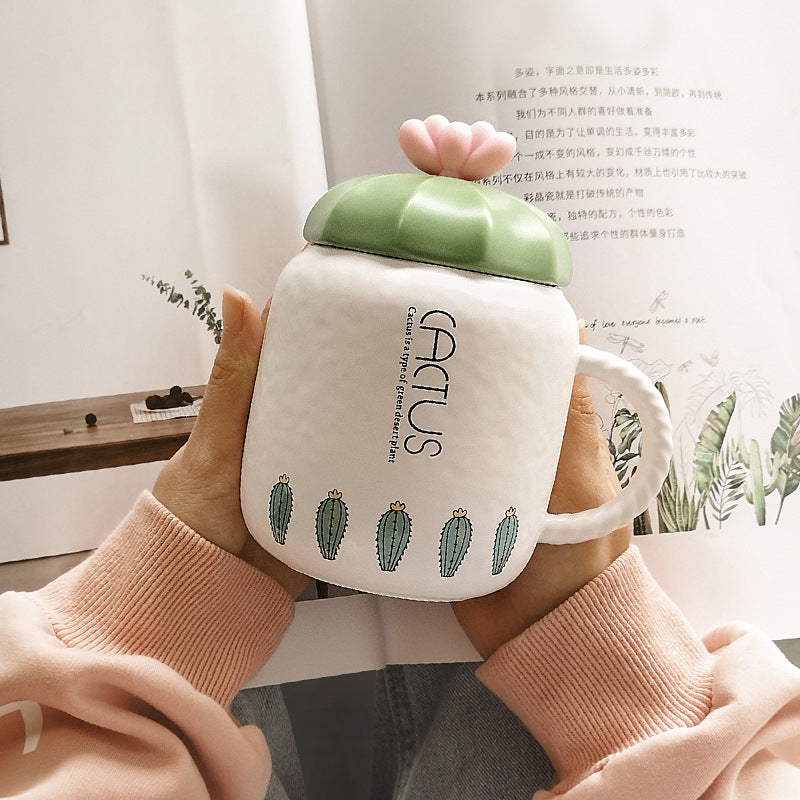 Person holding a white mug with green lid featuring cactus designs, labeled 'GENTUS'.