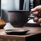 Hand holding a dark ceramic cup over a matching saucer on a wooden surface.