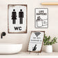 Three bathroom-themed signs on a wall with a sink and plant in the background.