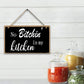 Decorative sign with text 'No Bitchin' in my kitchen' hanging on a white wooden wall with jars of spices below.