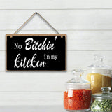 Decorative sign with text 'No Bitchin' in my kitchen' hanging on a white wooden wall with jars of spices below.