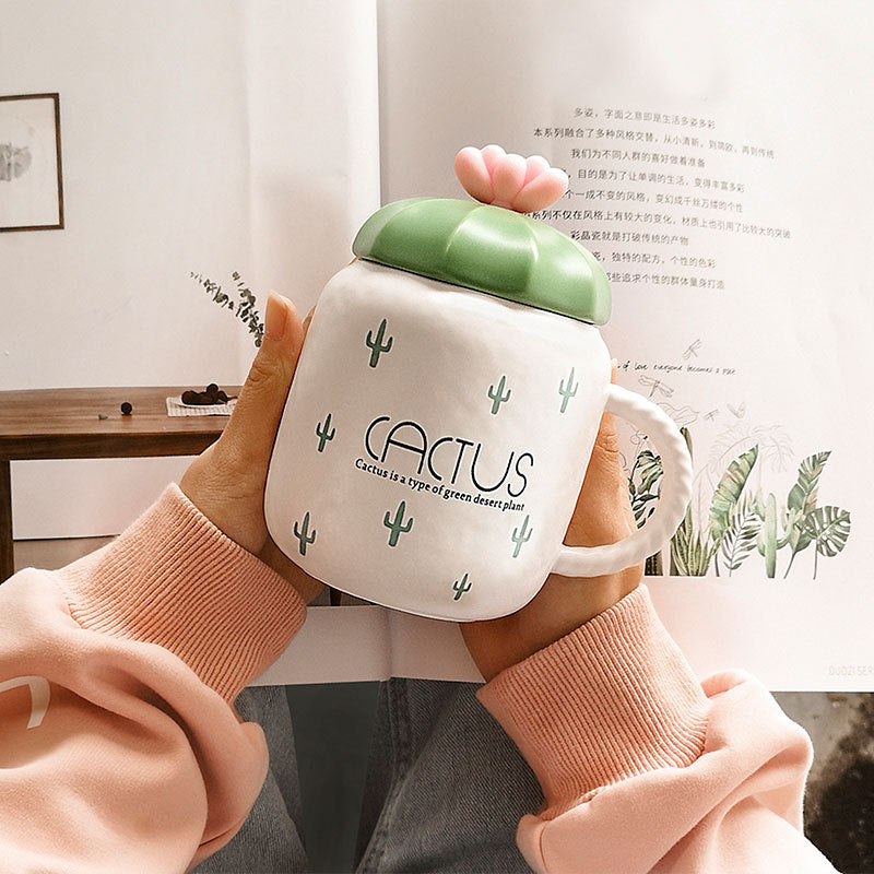 Person holding a white mug with green lid and cactus design, in front of a book.