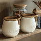 Two ceramic sugar jars with wooden lids and spoons on a shelf.