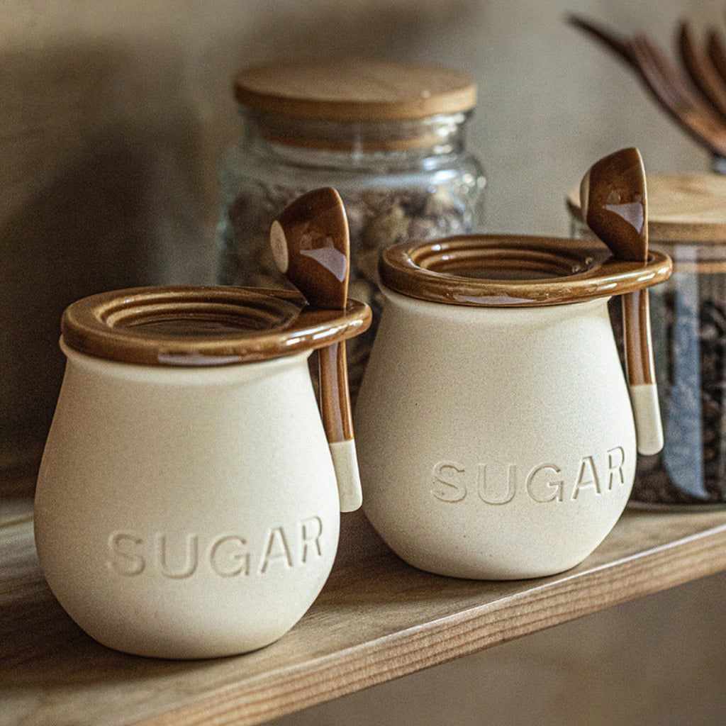 Two ceramic sugar jars with wooden lids and spoons on a shelf.