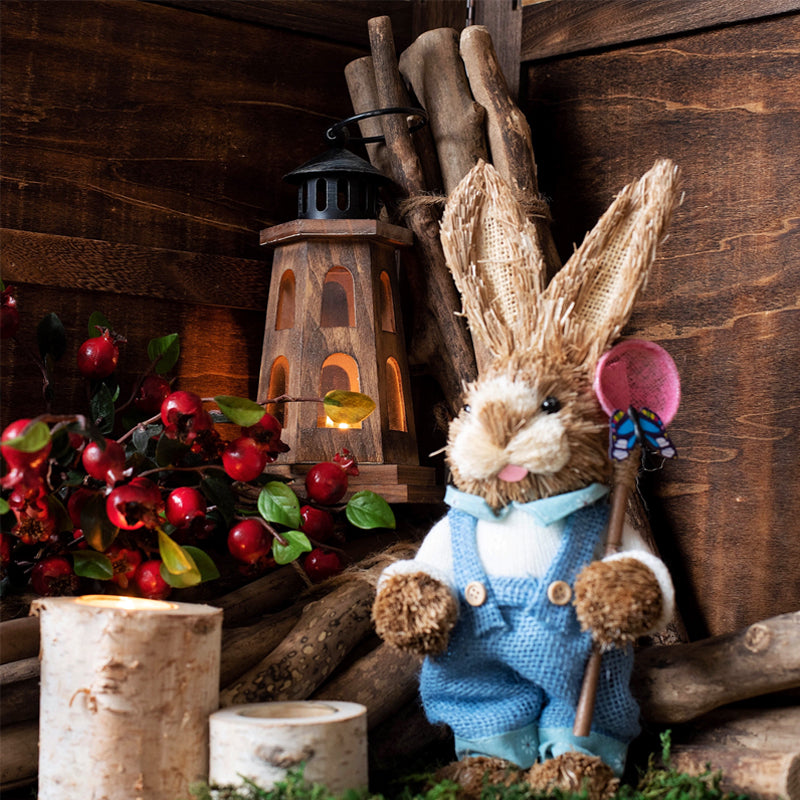Toy rabbit in overalls holding a pink flower in a rustic setting with logs and lantern.