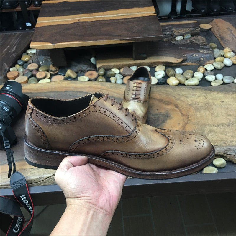 Brown leather dress shoe held by a hand on a wooden surface with pebbles.