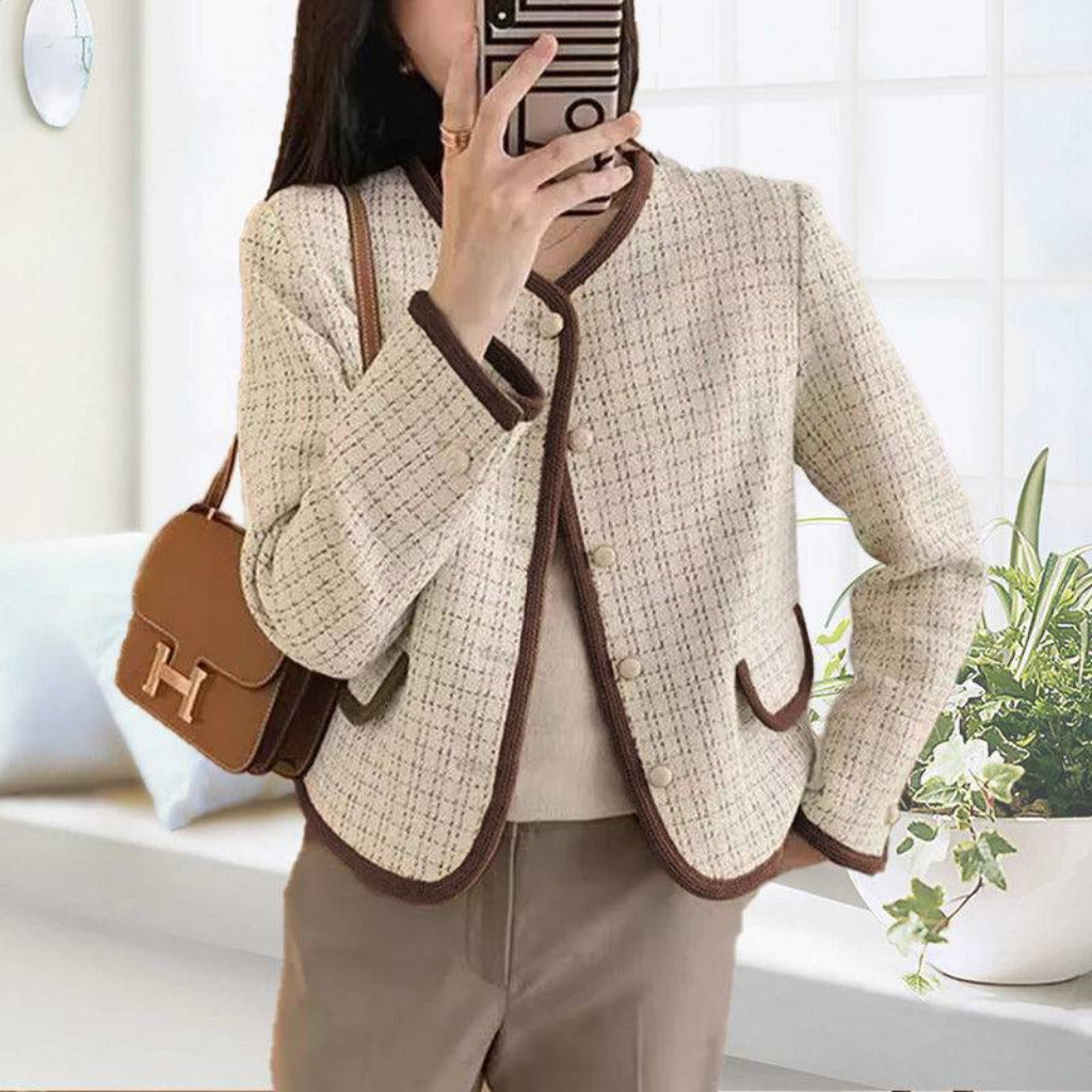 Person wearing a beige tweed jacket with brown trim, holding a phone, in a bright room.
