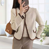 Person wearing a beige tweed jacket with brown trim, holding a phone, in a bright room.