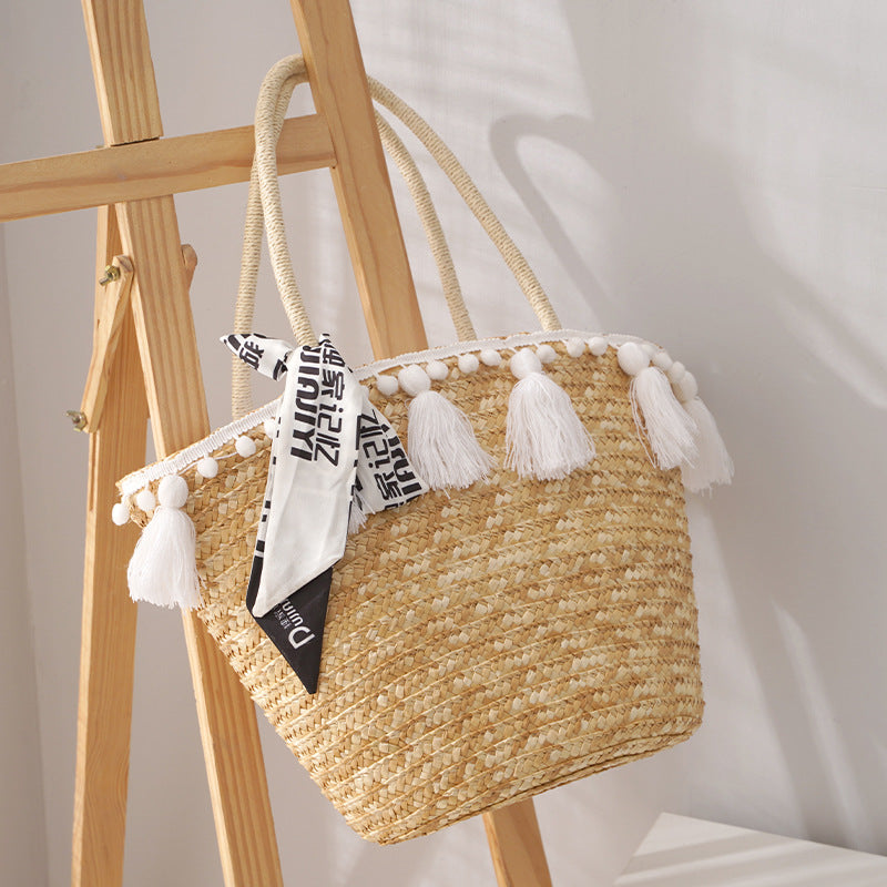 Woven straw bag with tassels on a wooden stand against a neutral background