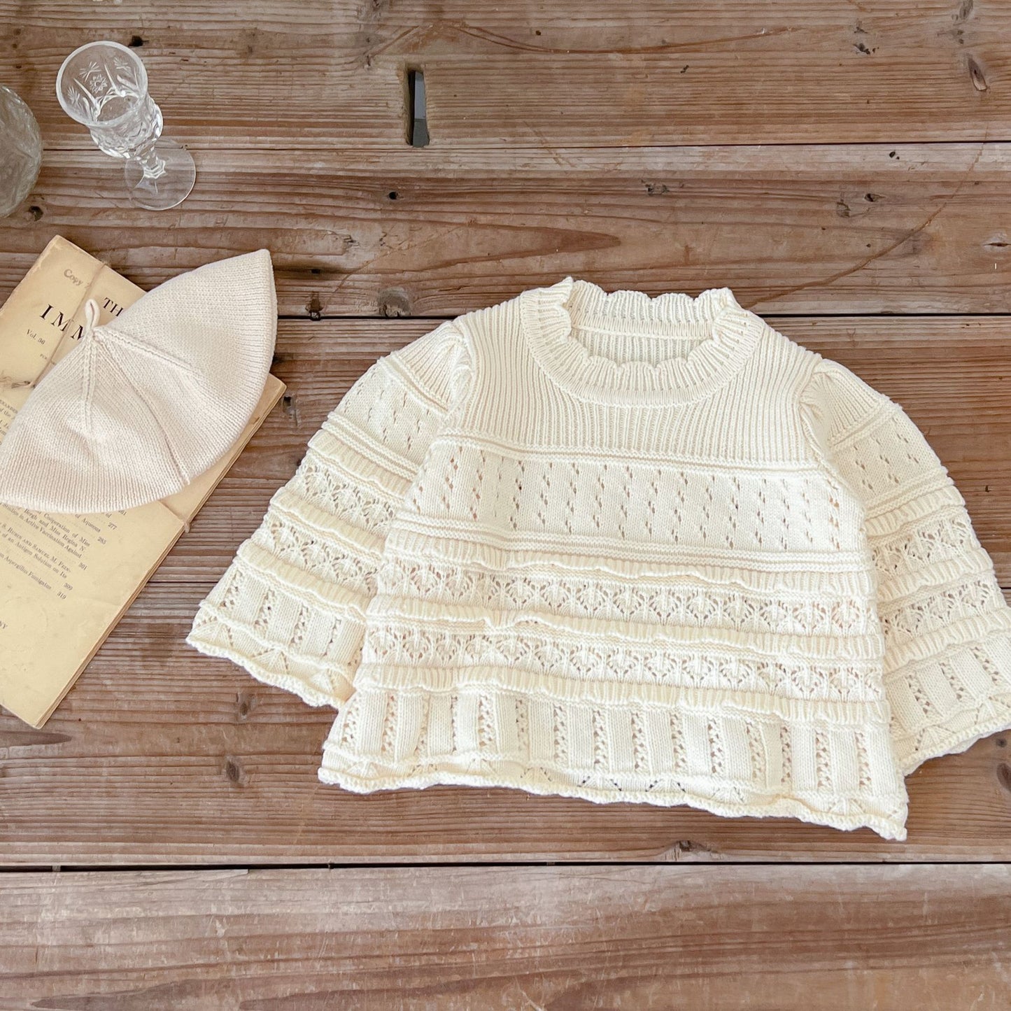 Knitted baby sweater and hat on a wooden surface with a glass of water and book.