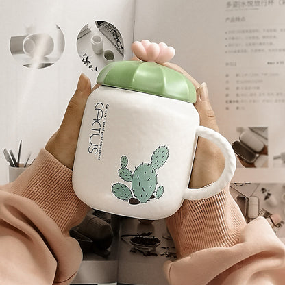 Hand holding a white mug with green lid and cactus design, against a blurred indoor background.