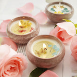 Three small candles with floral designs in ceramic holders surrounded by pink roses on a light wooden surface.