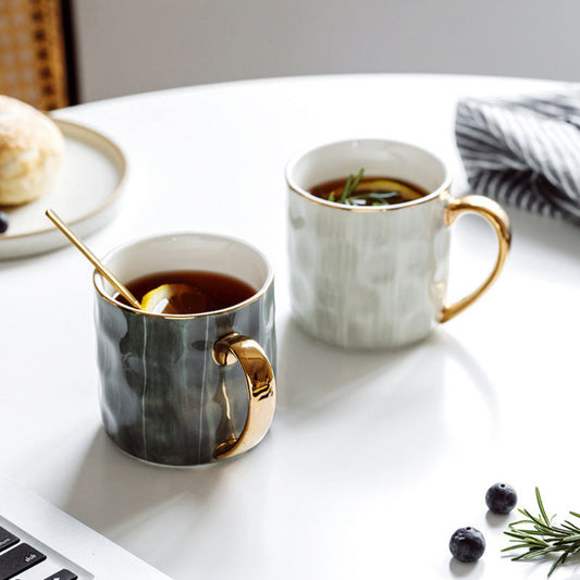 Two mugs with gold handles on a table next to a laptop, with a plate of food and a napkin in the background.