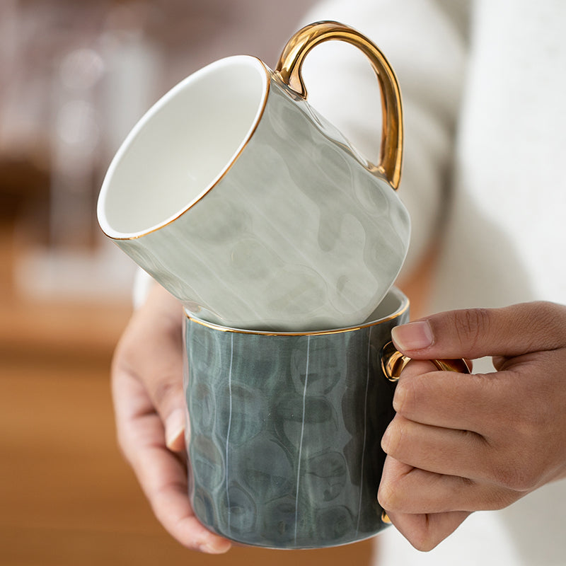 Two stacked ceramic mugs with gold handles held by a person.