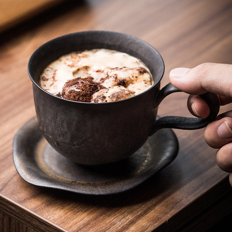 Person holding a dark ceramic mug with a hot chocolate drink on a wooden table