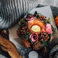 Decorative candle holder with flowers, pinecones, and a feather on a textured surface.