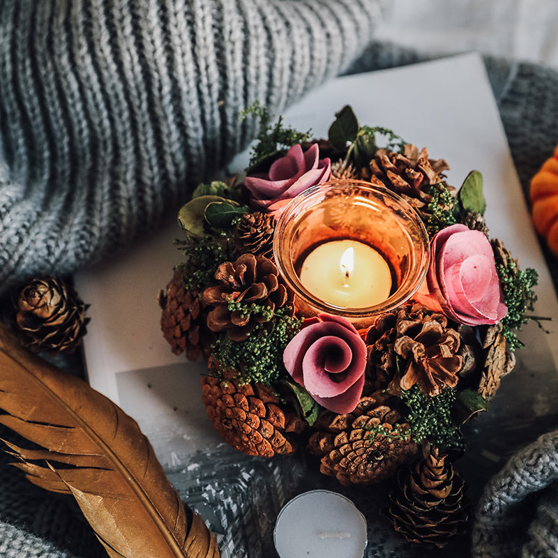 Decorative candle holder with flowers, pinecones, and a feather on a textured surface.