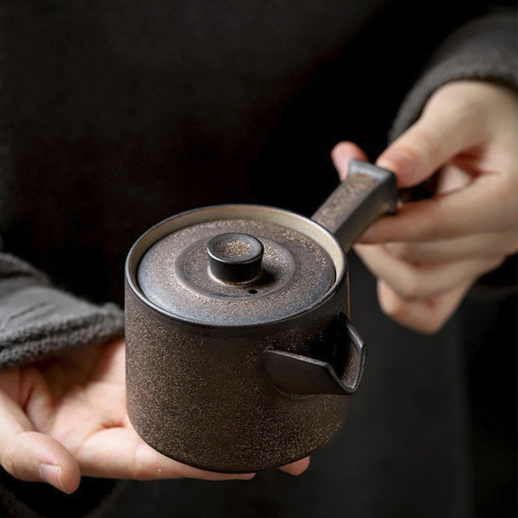 Person holding a black ceramic teapot with a lid and handle against a dark background