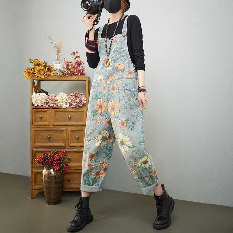 Person wearing floral overalls taking a mirror selfie in a room with a wooden dresser and decorative flowers.