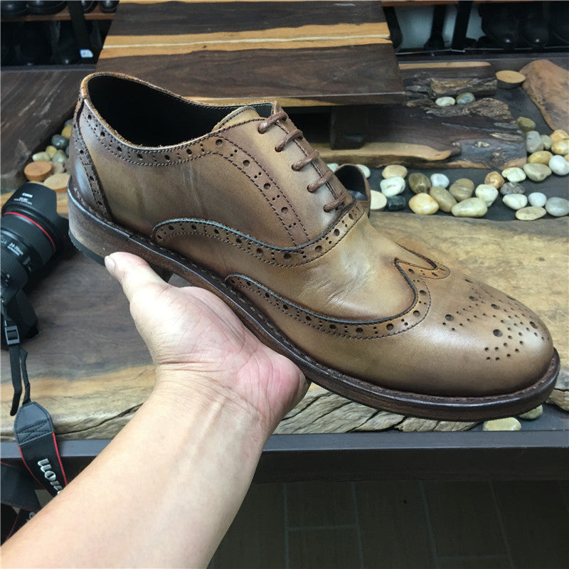 Brown leather wingtip shoe held by a hand on a wooden surface with pebbles.