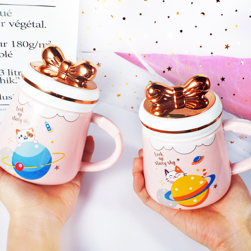 Two pink mugs with gold lids featuring cartoon designs held by hands against a white background.